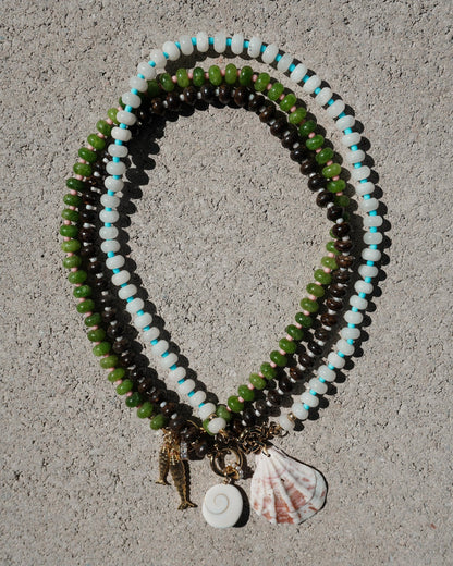 Three Mod + Jo Macey Gemstone Necklaces, arranged in ovals on a gray textured surface, feature genuine gemstones and pendants—a seahorse, spiral shell, and scallop shell. One design is accented with elegant white jade beads.