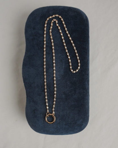 The Mod + Jo Callum Tennis Charm Clip Necklace, featuring a circular pendant and gold plating, is displayed atop a dark blue, velvet-textured case on a light surface.