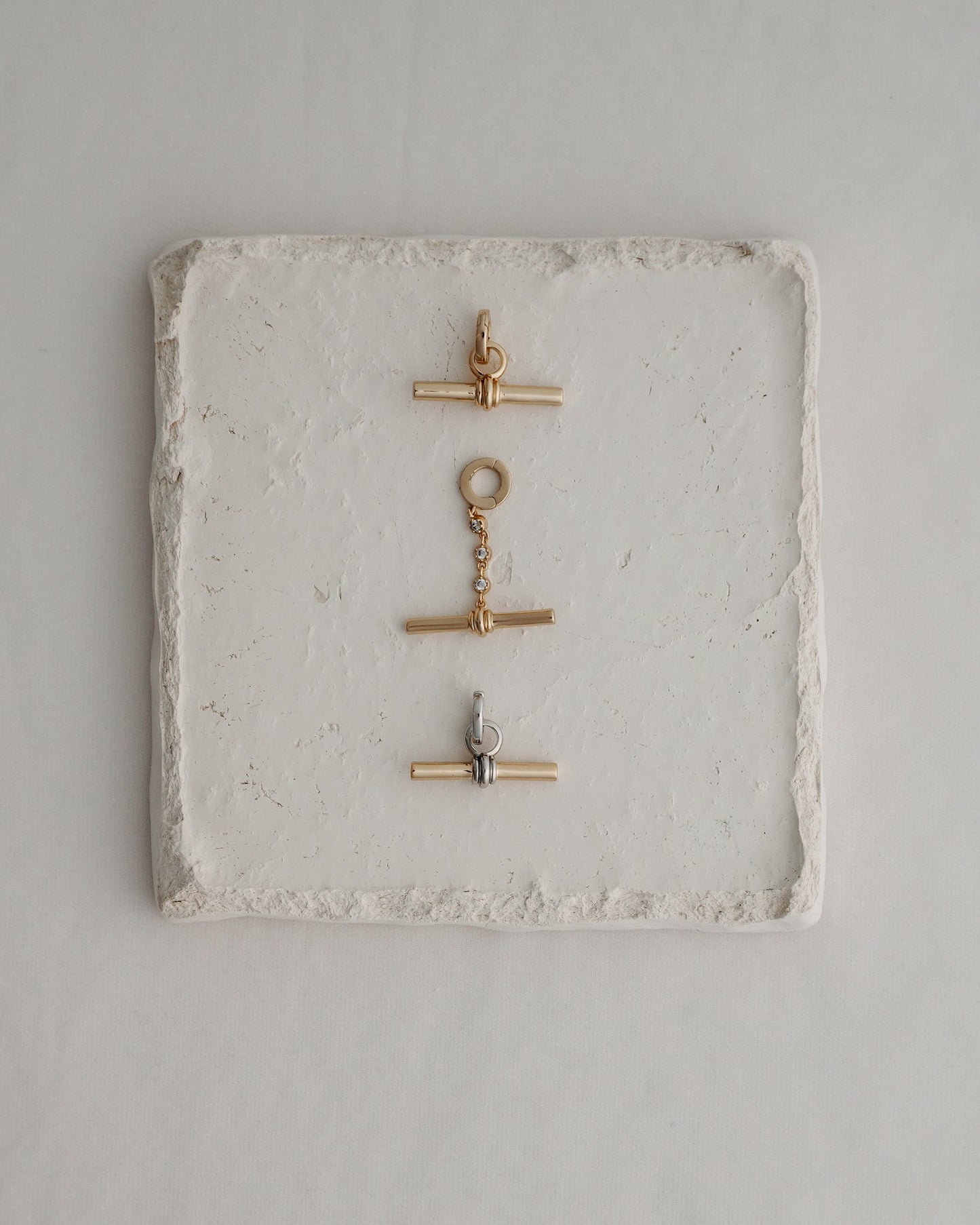 Three metal toggle clasps—two in gold and one in silver—are arranged vertically on a square, textured white stone tile set against a light background, echoing the elegant detail of Mod + Jo's Elliot Toggle Charm and the refined style characteristic of their 14k gold-inspired jewelry.