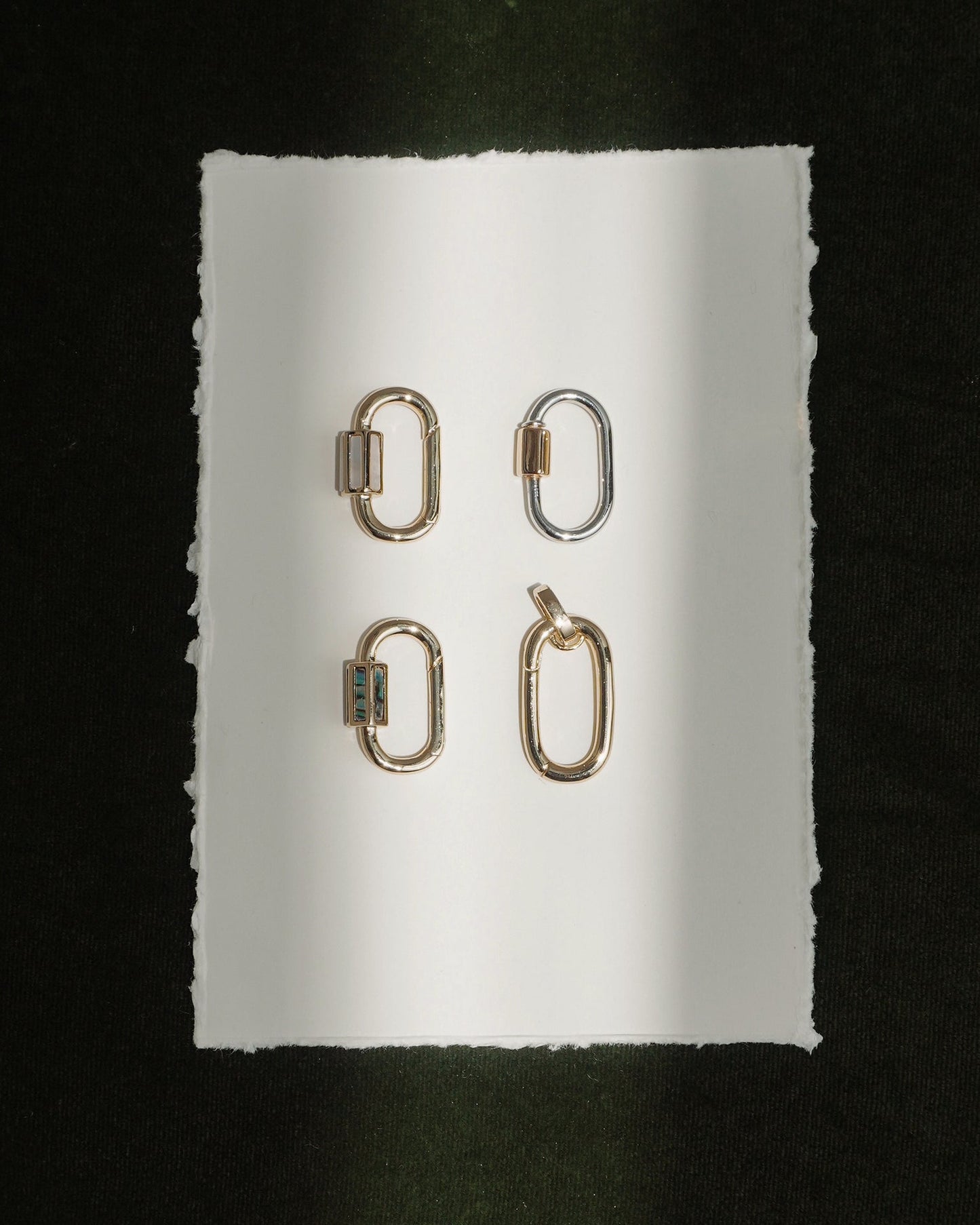 Four metallic Pip Charm Enhancers by Mod + Jo—three in radiant 14k gold and one in lustrous silver—are arranged in a grid on a white, rough-edged sheet of paper set against a dark background. A beam of light falls vertically across the arrangement, showcasing each enhancer like an elegant, modern charm accessory.