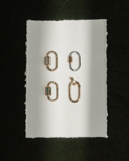 Four metallic Pip Charm Enhancers by Mod + Jo—three in radiant 14k gold and one in lustrous silver—are arranged in a grid on a white, rough-edged sheet of paper set against a dark background. A beam of light falls vertically across the arrangement, showcasing each enhancer like an elegant, modern charm accessory.