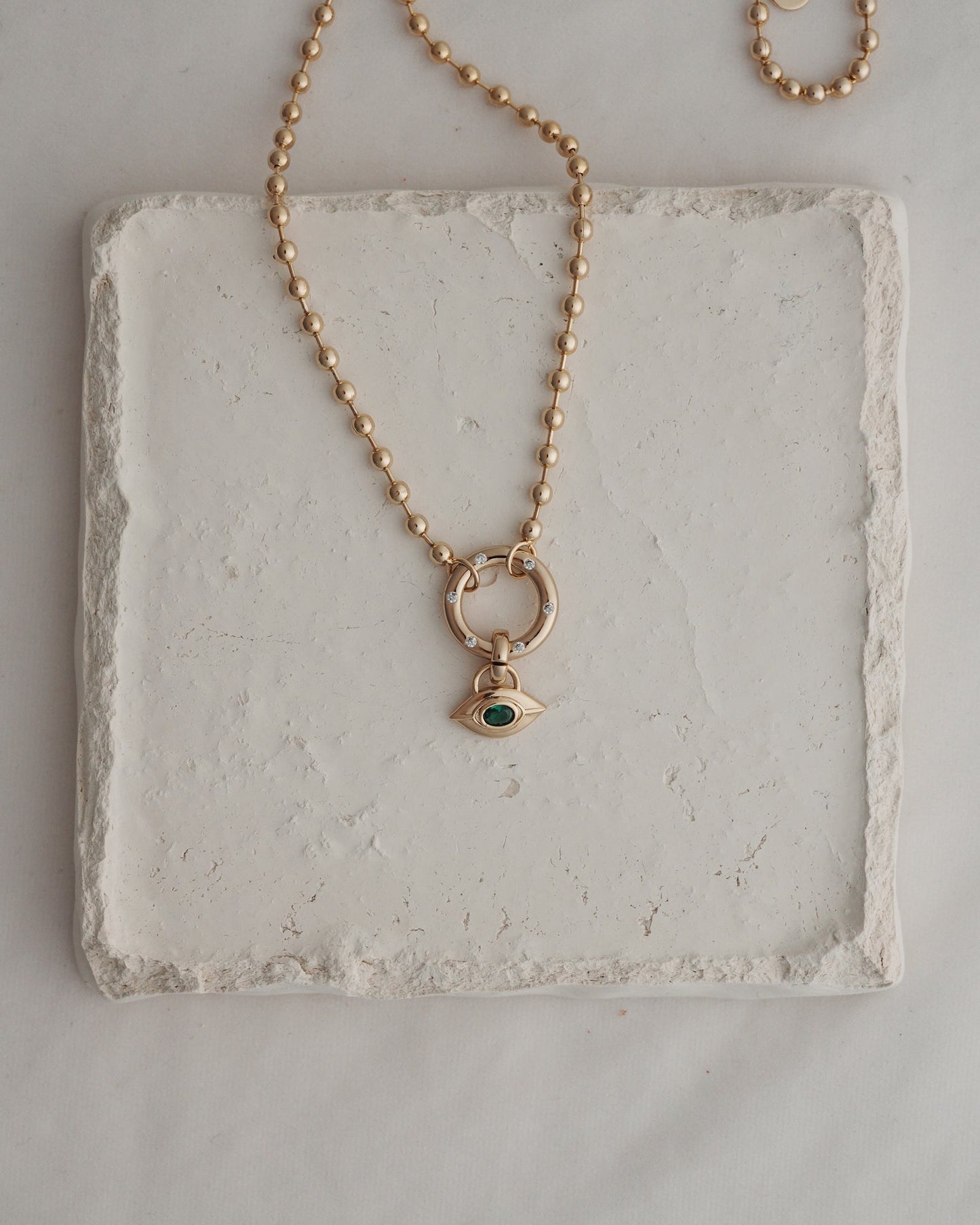 The Nyx Evil Eye Charm necklace by Mod + Jo features a round beaded gold chain and an eye-shaped pendant set with a green gemstone, artfully displayed on a textured white square tile—an elegantly crafted piece of protective jewelry that exudes the subtle allure of the evil eye.