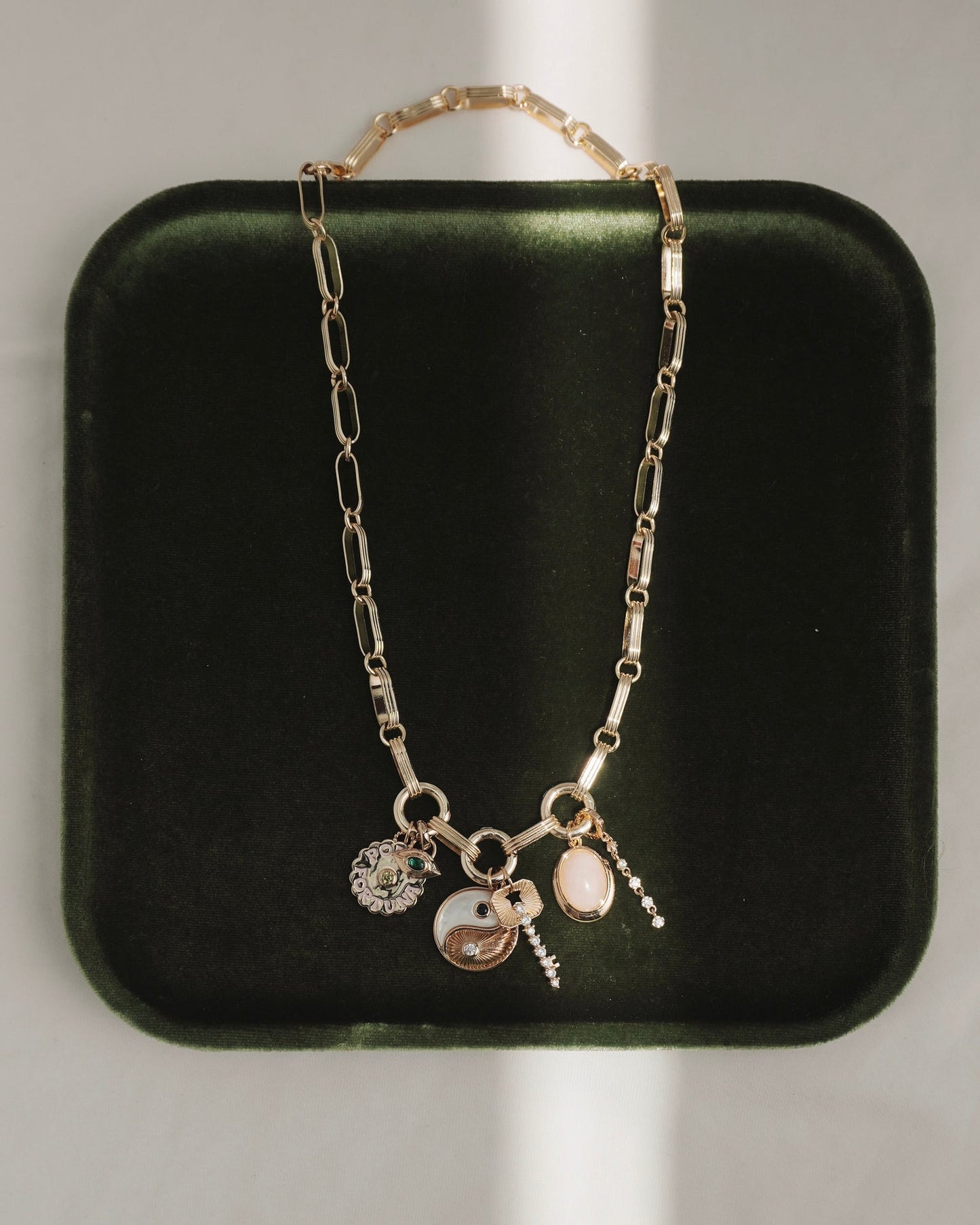 The Jagger Charm Clip Necklace by Mod + Jo, crafted in 14k gold and featuring four unique charms—a yin-yang, a flower, an oval stone, and a petite hanging pendant—is elegantly displayed on a dark green velvet tray with sunlight creating gentle shadows.