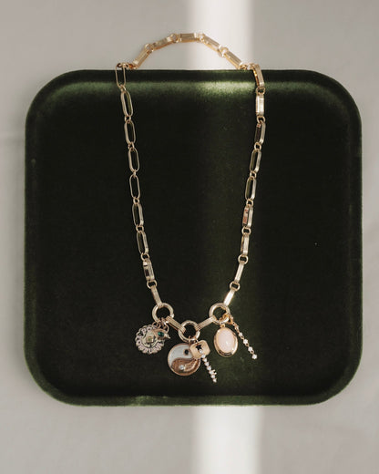 The Jagger Charm Clip Necklace by Mod + Jo, crafted in 14k gold and featuring four unique charms—a yin-yang, a flower, an oval stone, and a petite hanging pendant—is elegantly displayed on a dark green velvet tray with sunlight creating gentle shadows.