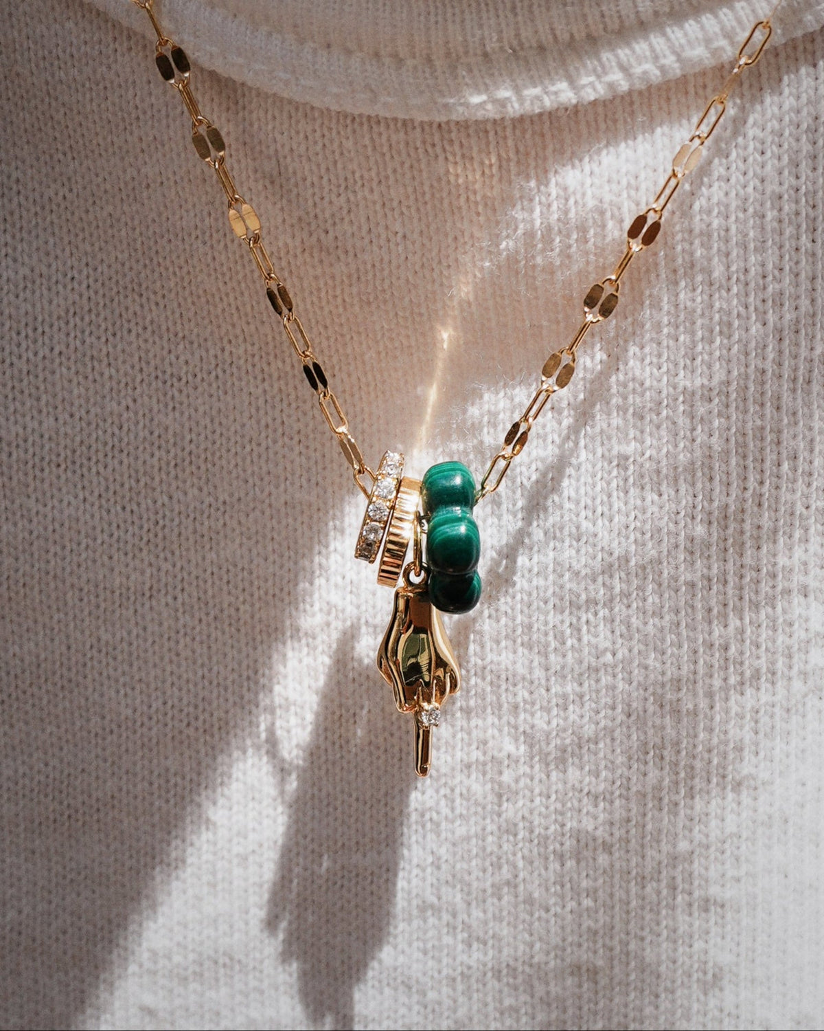 The Bird Charm by MOD + JO is a nickel-free 18k gold chain necklace with a bird pendant, crafted using advanced plating technology for lasting shine. Shown layered over textured cream fabric with green beads and a clear-stone ring charm.