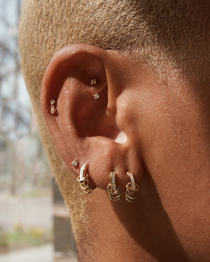 A close-up of an ear with several tarnish-resistant gold hoops, including Mod + Jo's Levi Huggies, and small mother of pearl studs. The wearer has short, light hair and is shown against a softly blurred outdoor background.