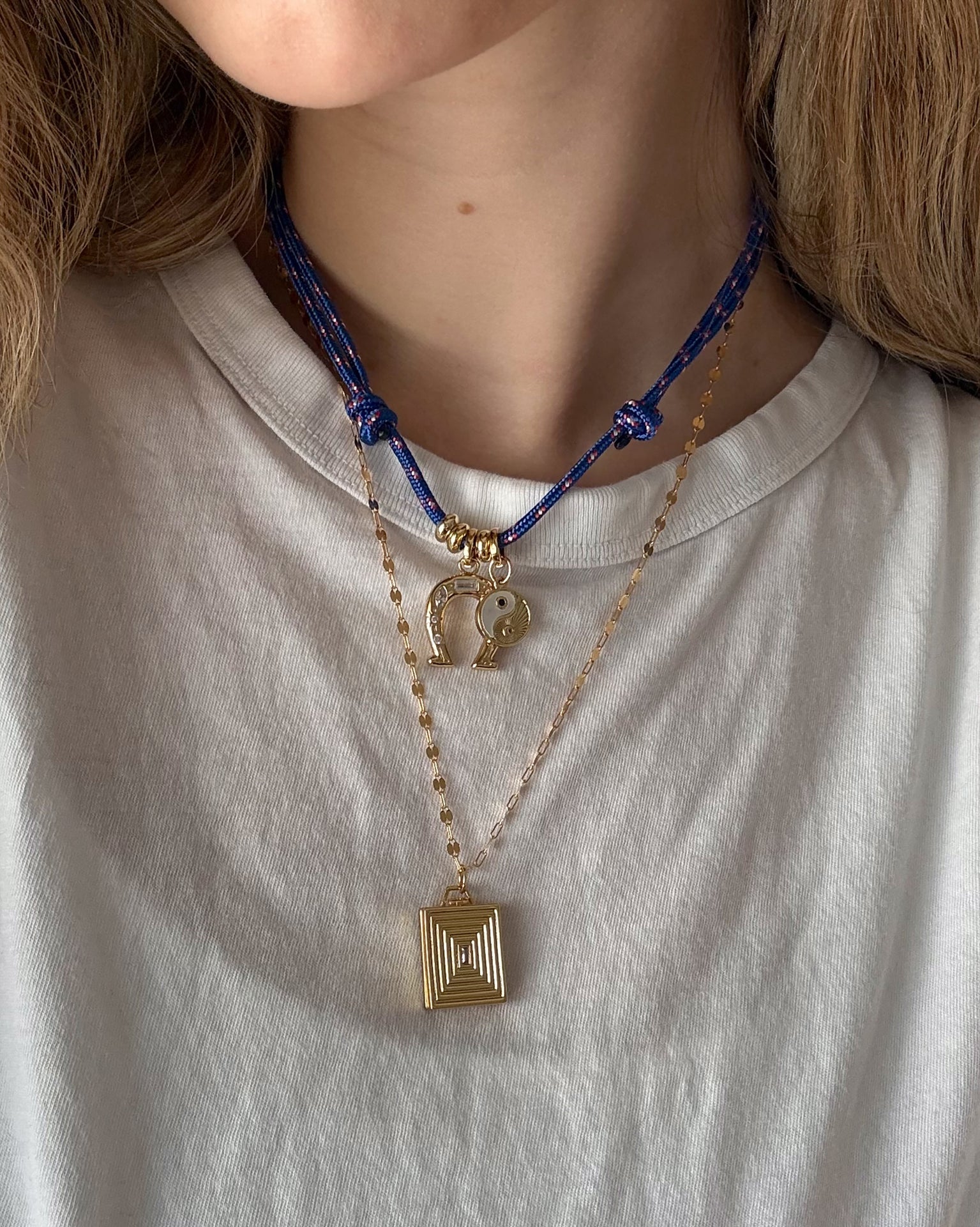 A person in a white t-shirt wears two necklaces: a bold blue cord necklace with gold charms, including a yin-yang, and Mod + Jo’s Inez Locket Charm—a delicate 14k gold plated chain with a square, textured pendant.