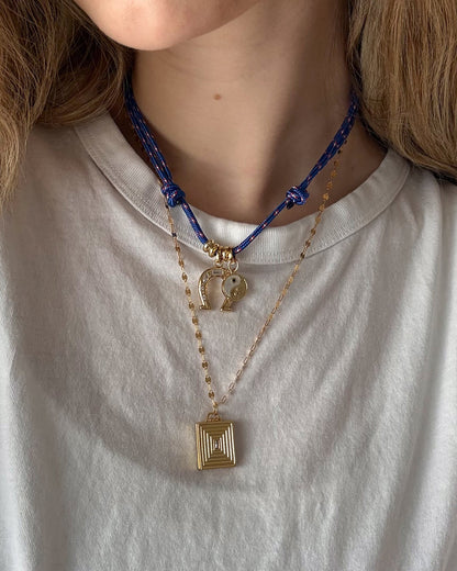 A person in a white t-shirt wears two necklaces: a bold blue cord necklace with gold charms, including a yin-yang, and Mod + Jo’s Inez Locket Charm—a delicate 14k gold plated chain with a square, textured pendant.