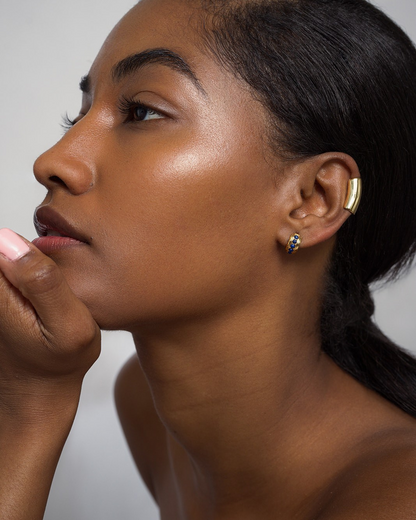 A woman with glowing skin and dark hair pulled back poses in profile, wearing Mod + Jo's Evy Raised Gemstone Hoops and a gold ear cuff. Her natural makeup and glossy lips highlight her elegant look as she lifts her hand near her chin.