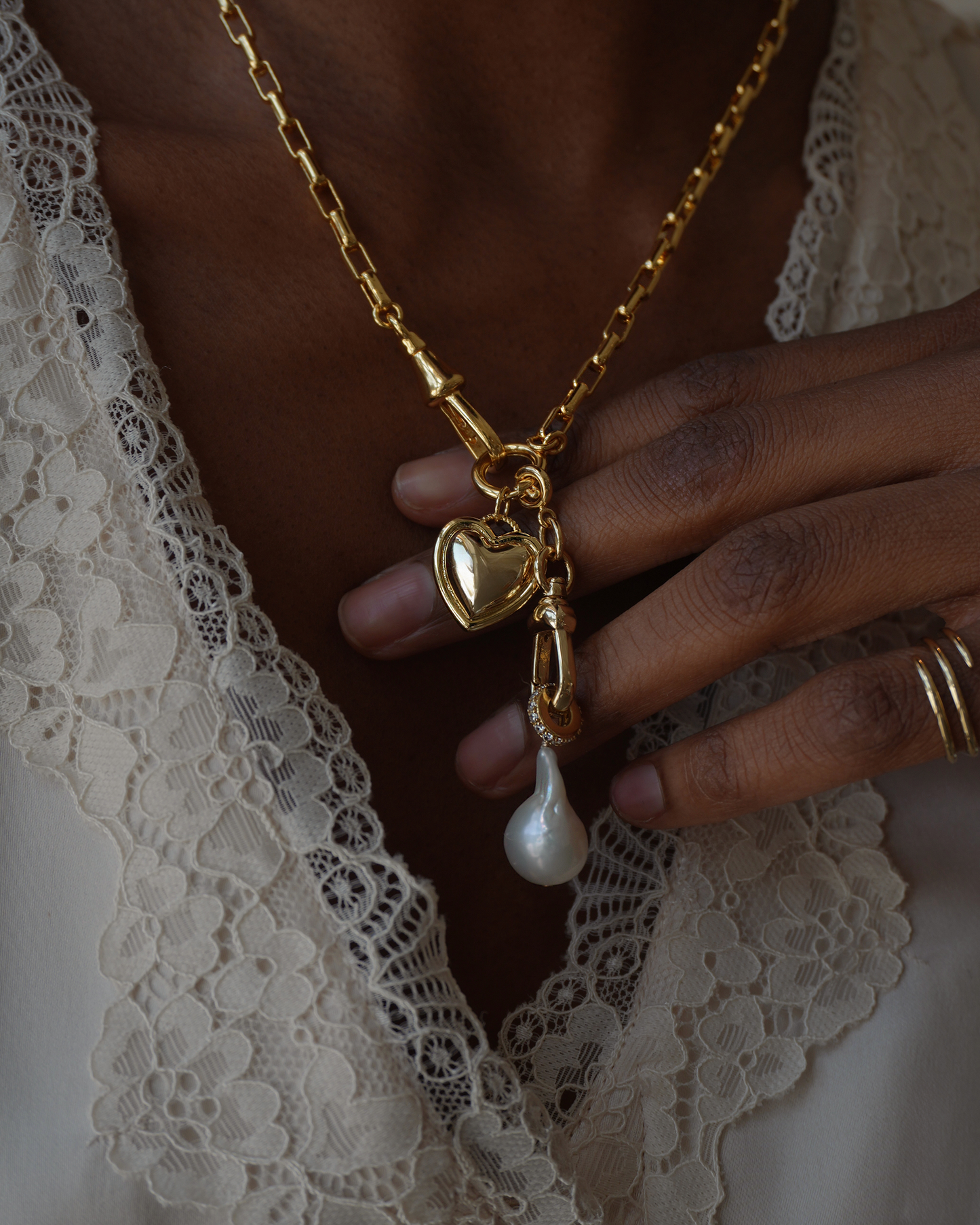 Close up of a gold link necklace with a heart pendant and pearl drop on women's neck wearing a white lace top