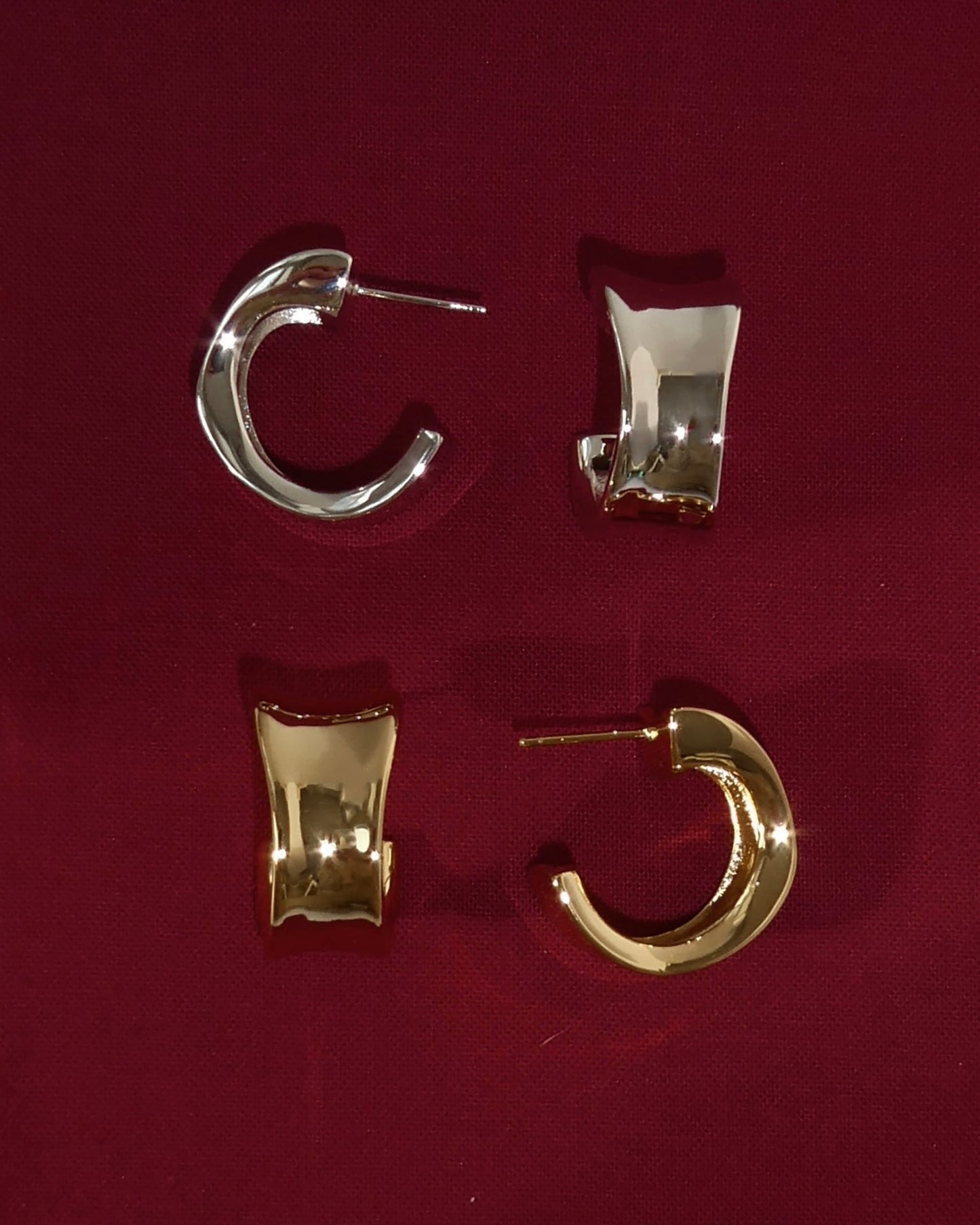Two pairs of Zara Volume Hoops by Mod + Jo—one chunky gold, one sleek silver—are displayed on dark red fabric. Both feature a modern, chunky design with smooth, shiny finishes.