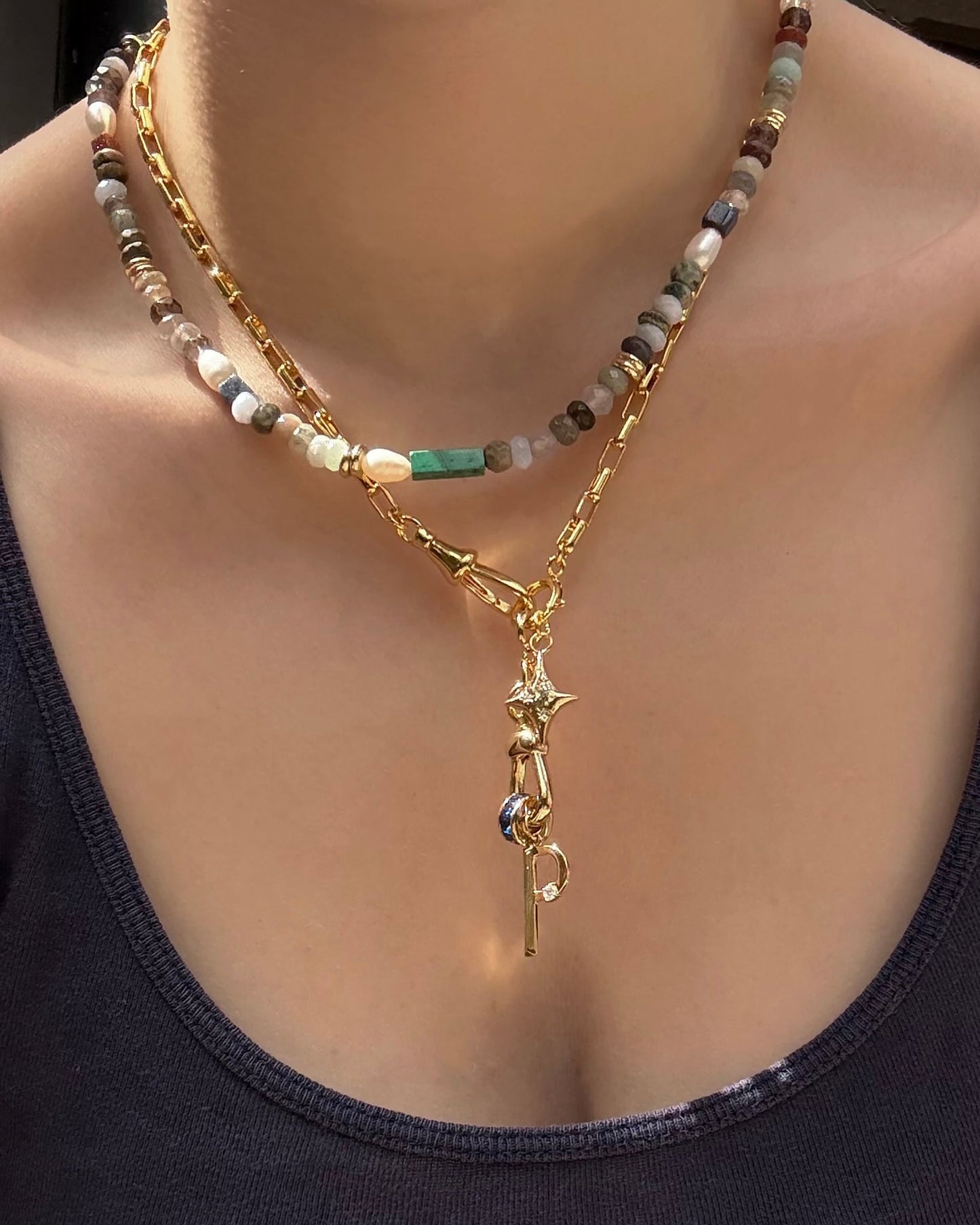 A person layers two necklaces over a dark tank: one with multicolored beads, the other—Mod + Jo’s Eloise Letter Charm—an 18k gold plated chain adorned with key, star, initial charm, and playful accents.