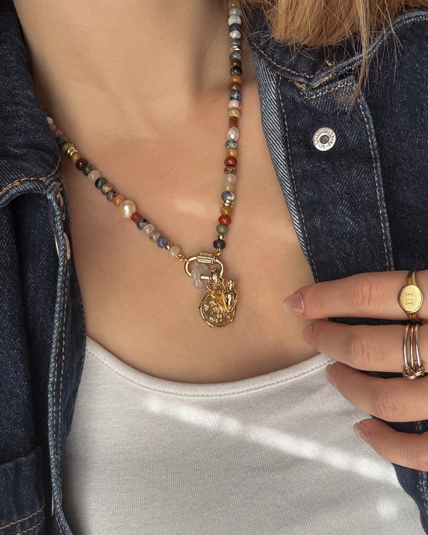 Someone wears Mod + Jo's Naomi Ancient Coin Charm beaded necklace with an 18k gold-plated charm, a denim jacket over a white top, and several gold rings.