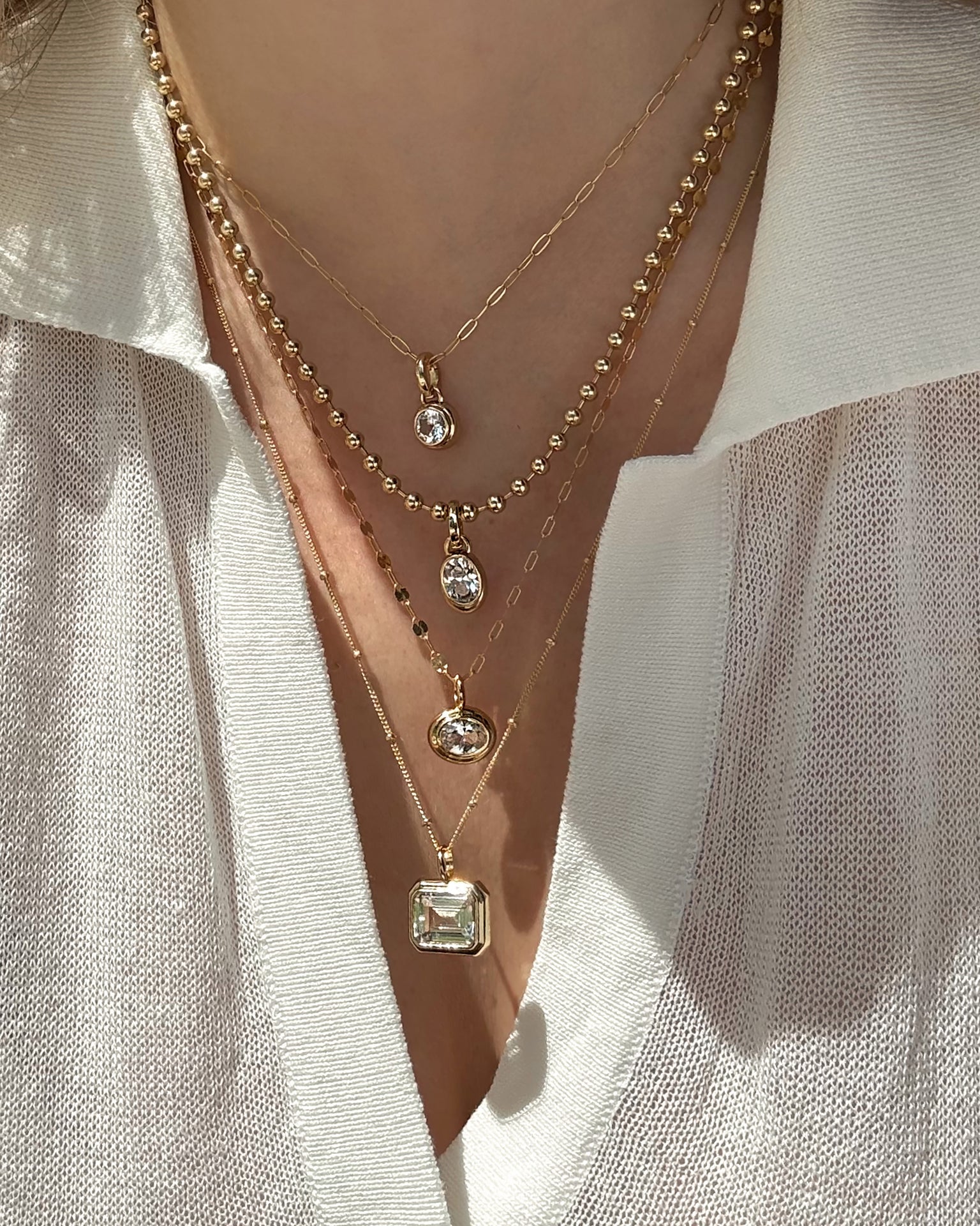 A person in a cream open-collar shirt displays four layered gold necklaces, including Mod + Jo's Elsie Sapphire Bezel Charm and a 14k gold pendant, each with lab-grown white sapphires in various shapes and sizes.
