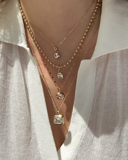 A person in a cream open-collar shirt displays four layered gold necklaces, including Mod + Jo's Elsie Sapphire Bezel Charm and a 14k gold pendant, each with lab-grown white sapphires in various shapes and sizes.