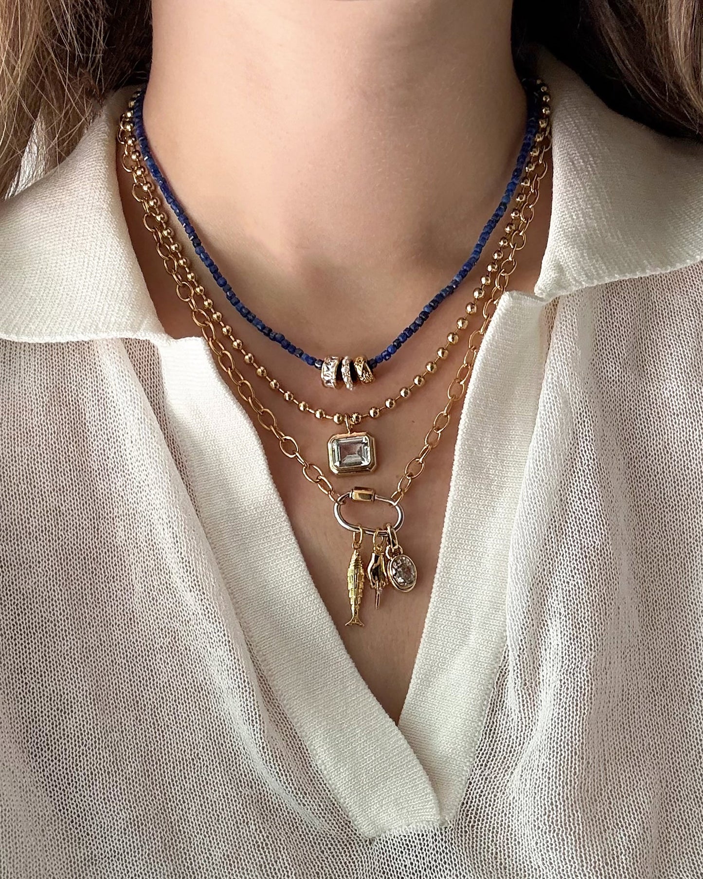 Close-up of a person wearing a white collared blouse with four layered necklaces, including Mod + Jo’s Nora Lock Carabiner: a blue beaded strand, two 14k gold-plated, tarnish-resistant chains with pendants, and a chain featuring three small rings and the signature Lock carabiner detail.