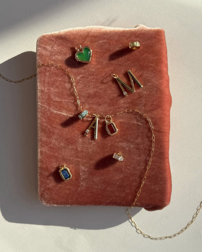 Mod + Jo’s Este Birthstone Charm Spacer, gold plated necklace, personalized letter pendants A and M, and colored gemstone jewelry are displayed on a soft pink velvet surface in sunlight.