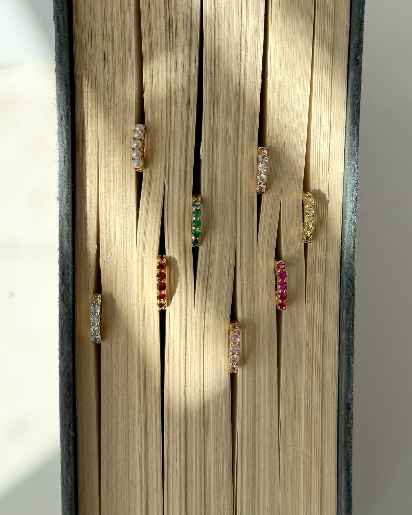The Mod + Jo Harlow Reversible Birthstone Huggies are displayed between the pages of an open book, with soft natural light casting gentle shadows on the pages.