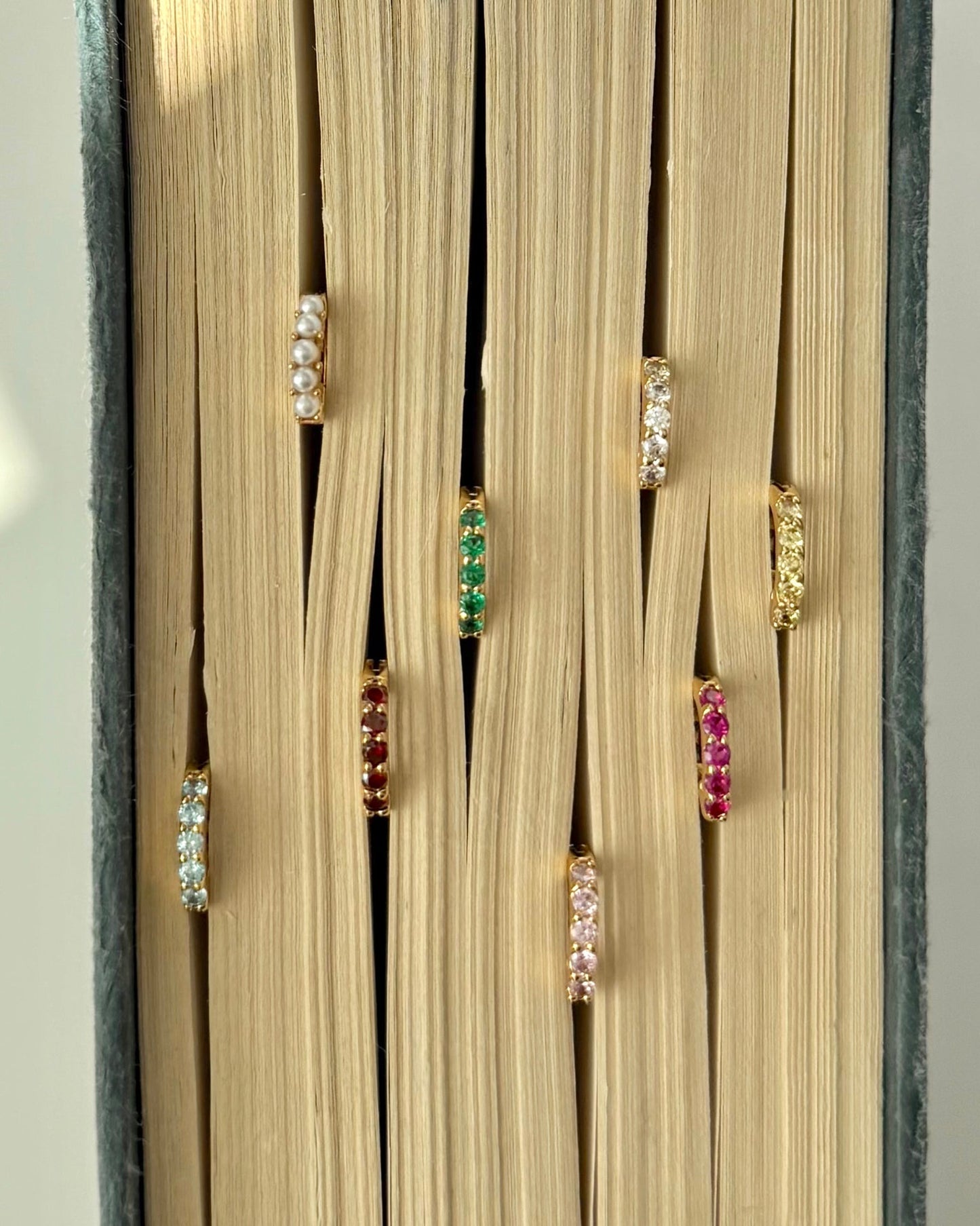 Seven pairs of Mod + Jo Harlow Reversible Birthstone Huggies, each featuring a different colored gemstone, are shown inserted between the closed pages of a book, with the birthstone earrings peeking out.