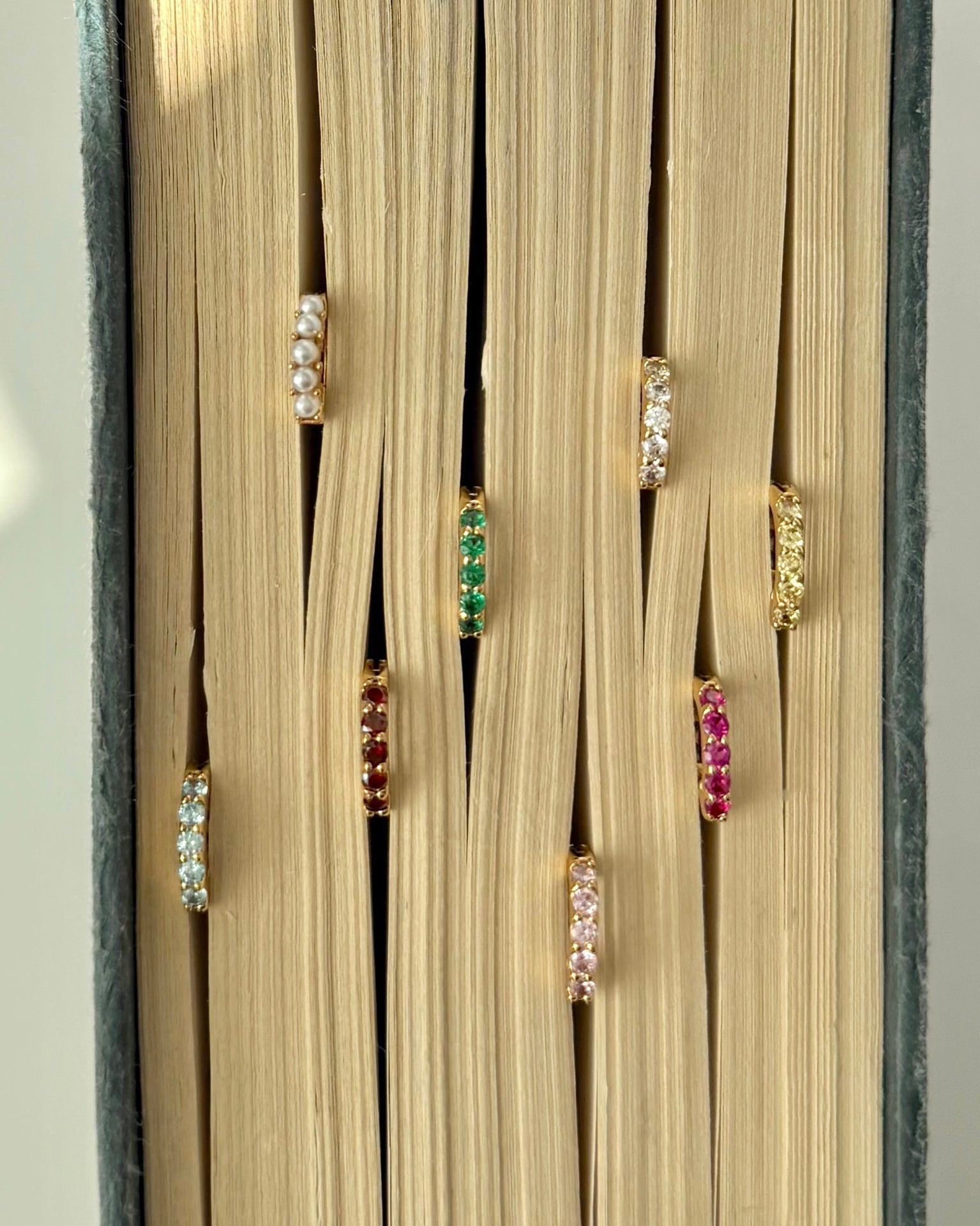 Seven pairs of Mod + Jo Harlow Reversible Birthstone Huggies, each featuring a different colored gemstone, are shown inserted between the closed pages of a book, with the birthstone earrings peeking out.
