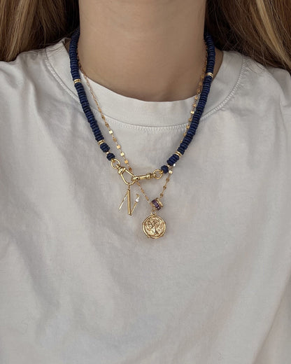 A person wears a white shirt with two layered necklaces: Mod + Jo's Eloise Letter Charm—a personalized gold initial and medallion—and a second necklace with dark blue beads and gold accents.