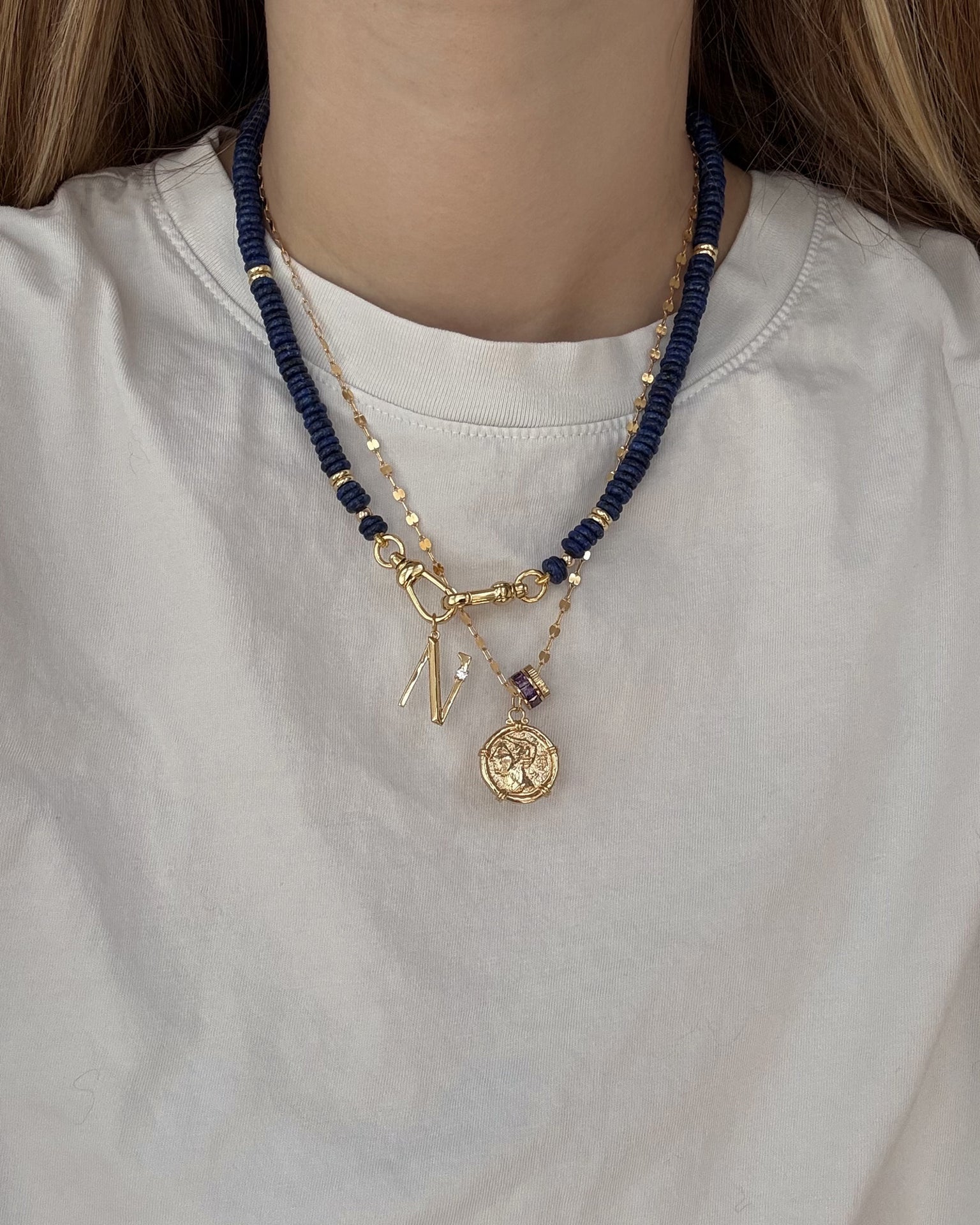 A person wearing a white shirt with three layered necklaces: one gold plated letter charm necklace, one with dark blue beads, and the Mod + Jo Este Birthstone Charm Spacer—a chic blend of personalized accessories.
