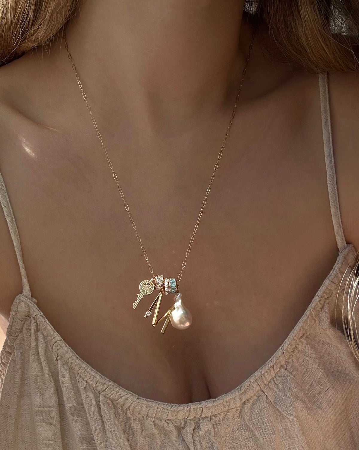 A woman wears a light beige spaghetti strap top and the Mod + Jo Frankie Key Charm gold necklace, featuring an Italian engraved key, a crystal letter “M,” a tassel, a pearl, and a small blue gem.