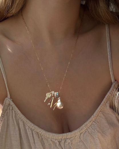 A woman wears a light beige spaghetti strap top and the Mod + Jo Frankie Key Charm gold necklace, featuring an Italian engraved key, a crystal letter “M,” a tassel, a pearl, and a small blue gem.