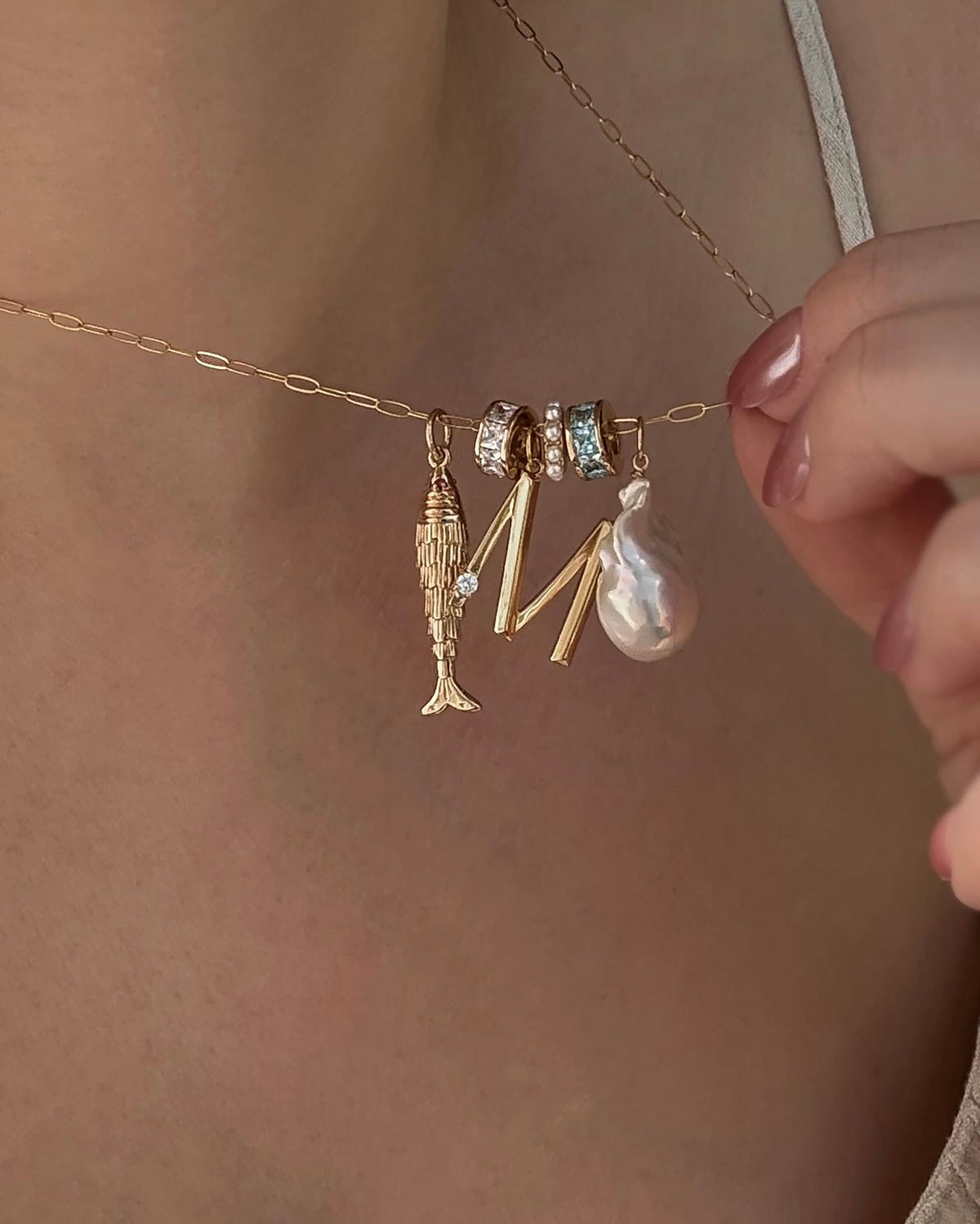 A close-up of the Mod + Jo Shia Fish Charm 14k gold-plated necklace, with two gemstone-studded rings, a gold letter M, and a pearl pendant, held by someone with light pink nail polish.