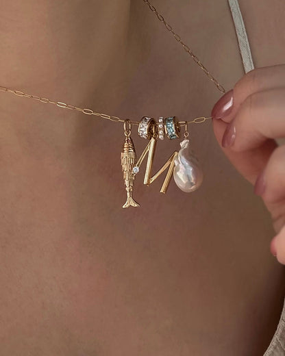 A close-up of the Mod + Jo Shia Fish Charm 14k gold-plated necklace, with two gemstone-studded rings, a gold letter M, and a pearl pendant, held by someone with light pink nail polish.