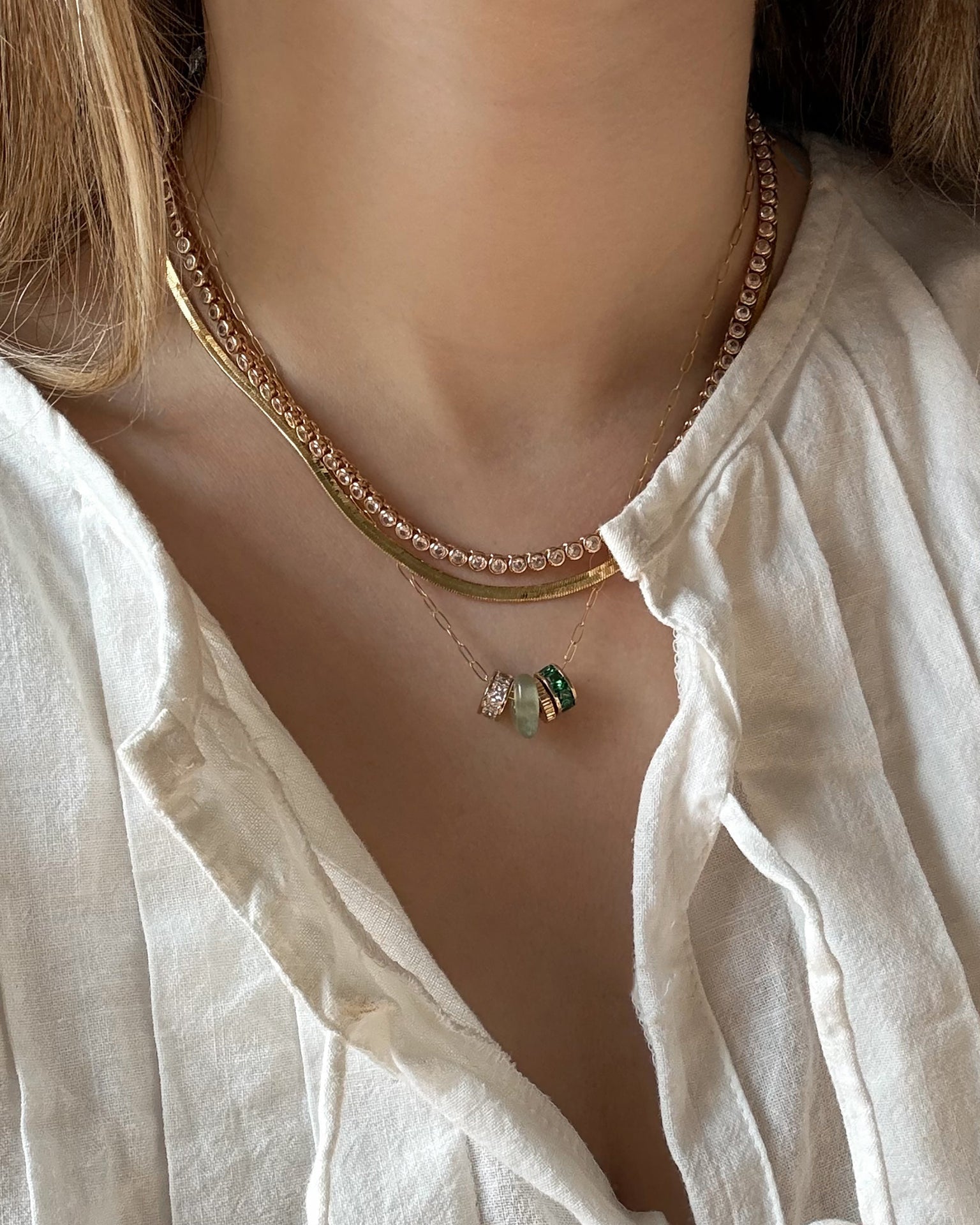 A person wearing a cream ruffled blouse and three layered gold necklaces, including Mod + Jo's Este Birthstone Charm Spacer, personalized beaded details, pendants with green and gold accents for a distinctive jewelry look.