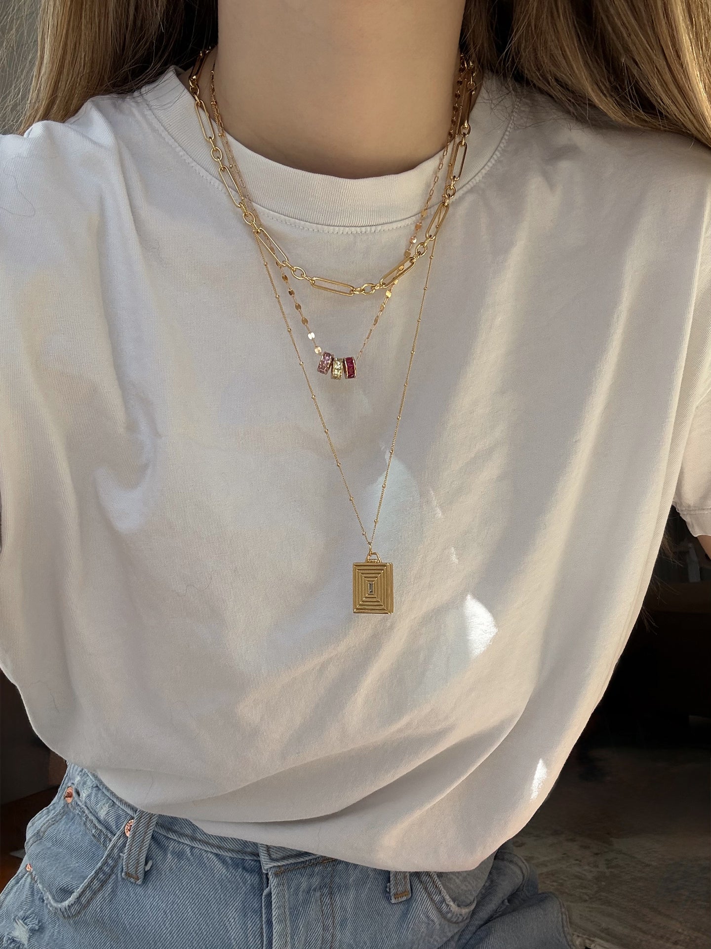 A person in a white tee and light blue jeans, layered with three gold necklaces—including Mod + Jo’s Inez Locket Charm, featuring a 14k gold plated rectangular pendant—plus colorful beads and a simple chain. Their long hair falls over their shoulders.