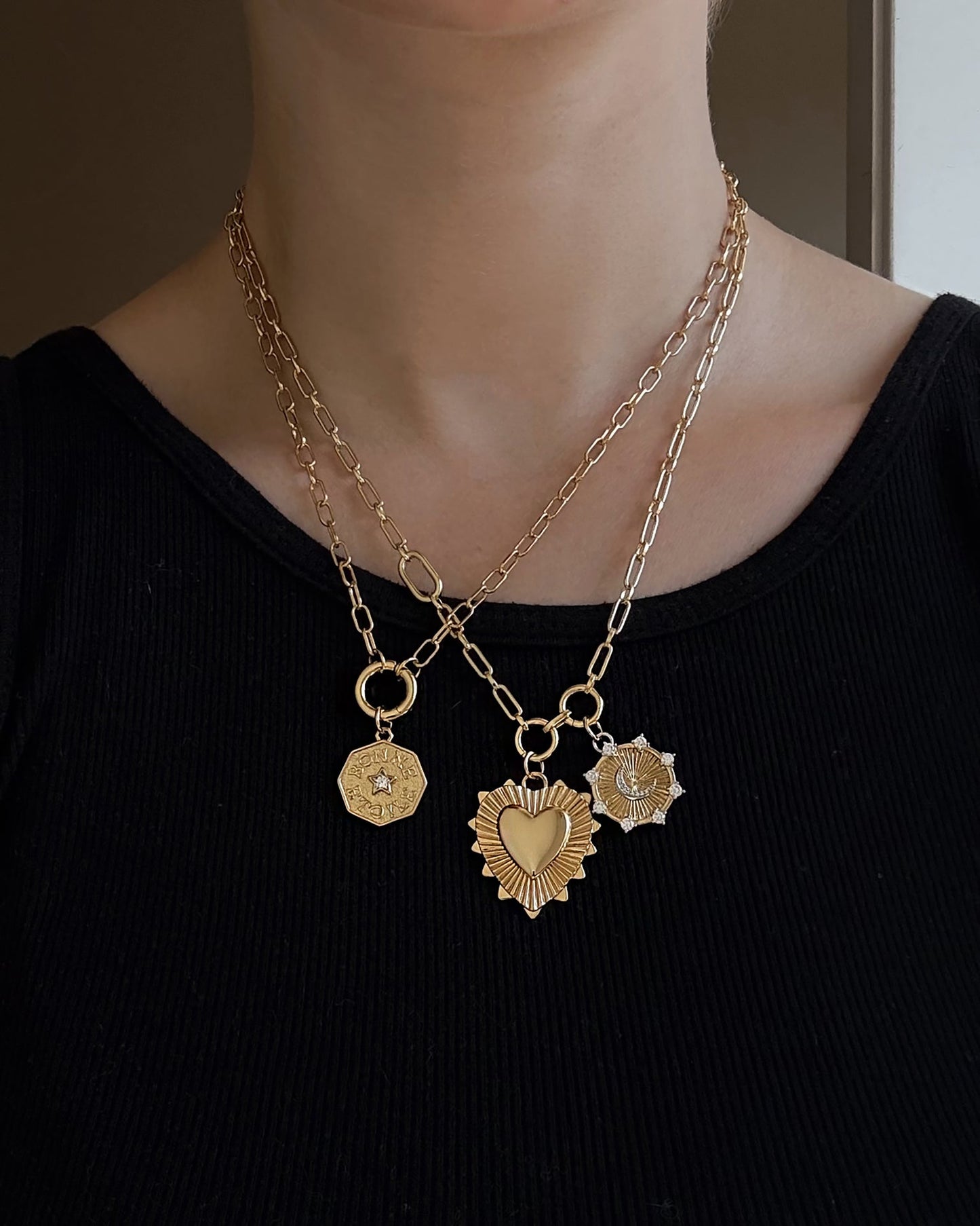 A person wears a black top and two gold chain necklaces adorned with three gold pendants—a hexagon, a radiant heart, and the Mod + Jo Bonne Etoile Charm featuring a sunburst with a crescent moon and stars—elegantly styled to evoke the timeless allure of gold coin jewelry while adding a touch of celestial luck and beauty.