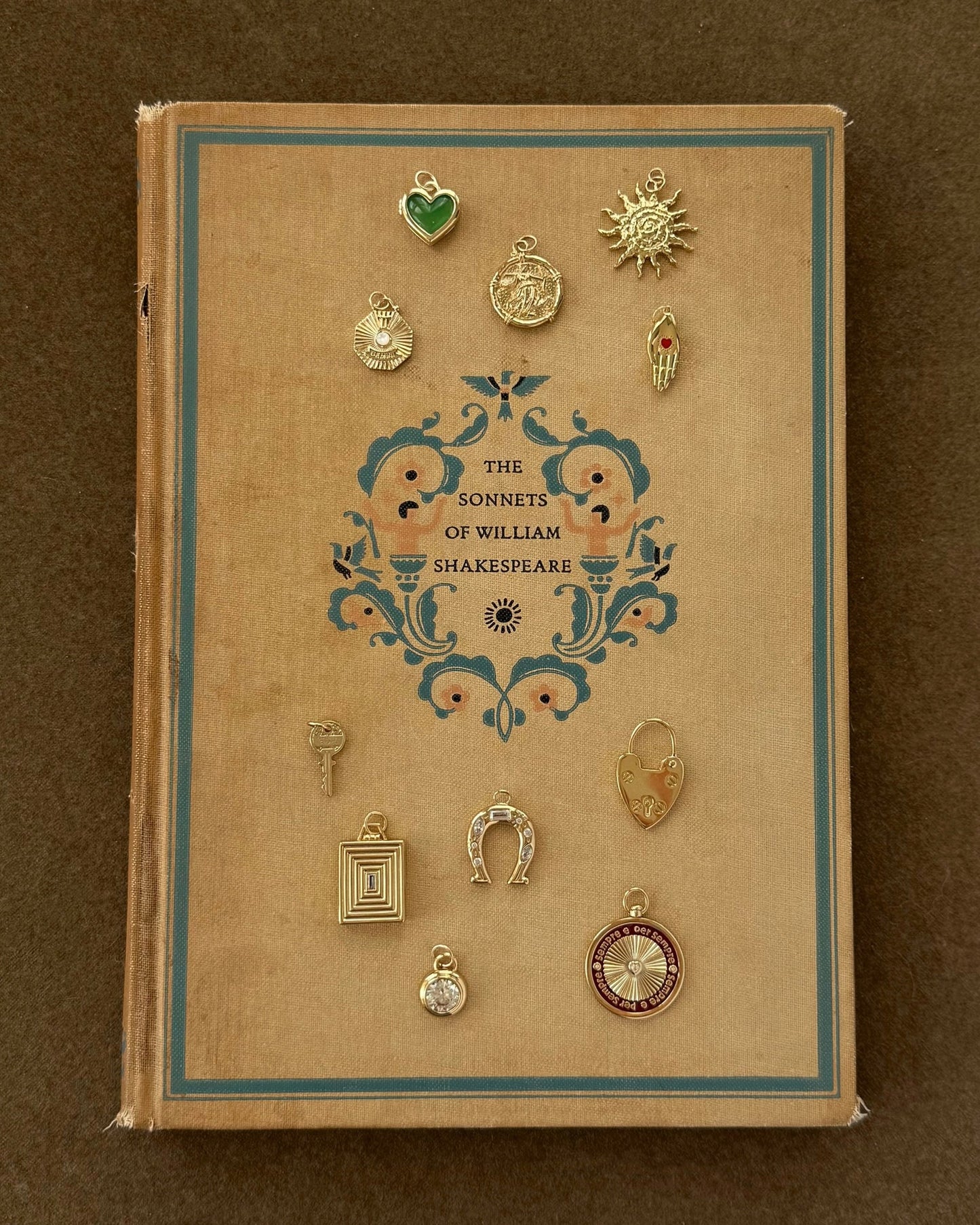 A vintage hardcover of The Sonnets of William Shakespeare sits on a brown surface, topped with twelve small 14k gold charms, including the Sasha Heart Locket Enhancer by Mod + Jo, a horseshoe, compass, and sun.