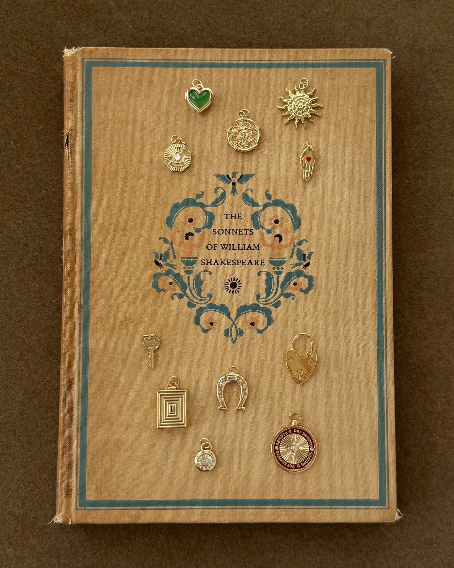 A vintage hardcover of The Sonnets of William Shakespeare sits on a brown surface, topped with twelve small 14k gold charms, including the Sasha Heart Locket Enhancer by Mod + Jo, a horseshoe, compass, and sun.