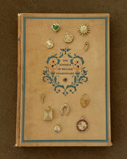 A vintage hardcover of The Sonnets of William Shakespeare sits on a brown surface, topped with twelve small 14k gold charms, including the Sasha Heart Locket Enhancer by Mod + Jo, a horseshoe, compass, and sun.