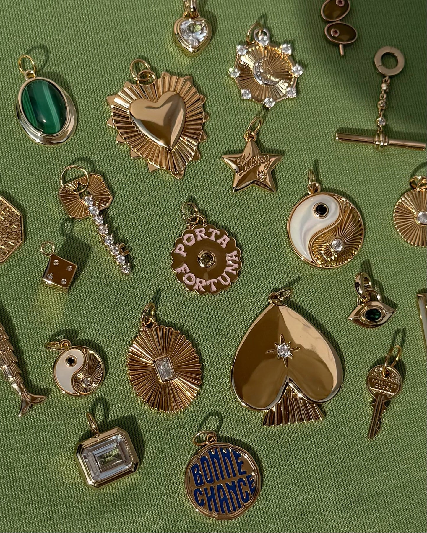 Assorted gold and gemstone charms, including the Mod + Jo Grande Lynn Engravable Heart Charm in 14k gold over brass, as well as stars, yin-yang symbols, dice, and a spade, are arranged on a textured green fabric background.