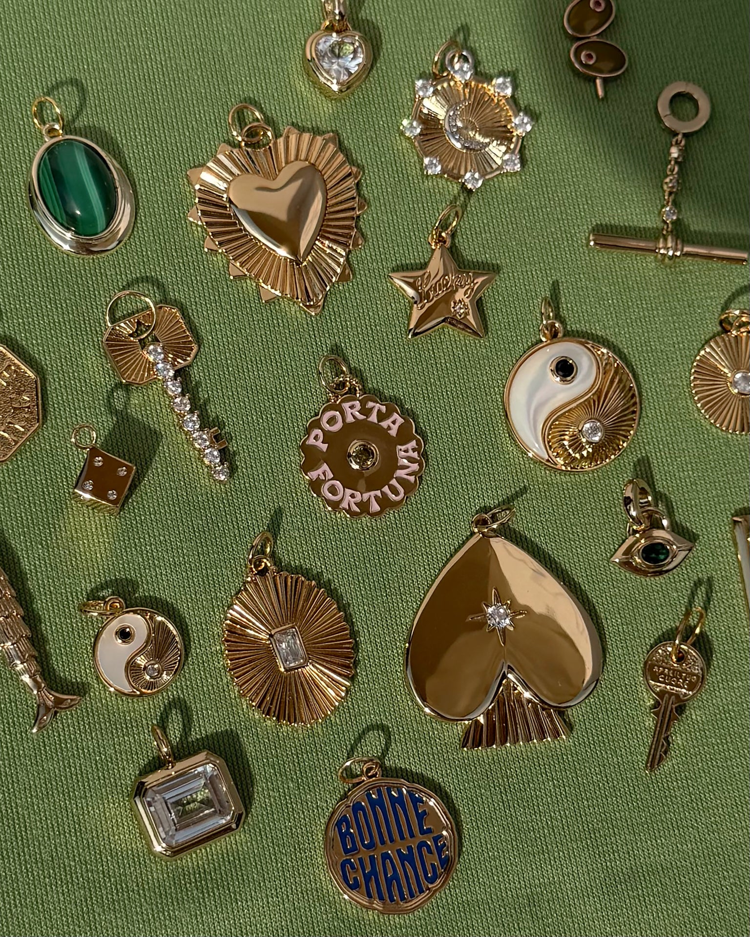 Assorted gold and gemstone charms, including the Mod + Jo Grande Lynn Engravable Heart Charm in 14k gold over brass, as well as stars, yin-yang symbols, dice, and a spade, are arranged on a textured green fabric background.