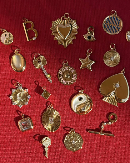 Assorted Mod + Jo 14k gold charms, including hearts, stars, yin-yang symbols, letters, and circular tokens, are displayed alongside the Veya Spade Charm and a sparkling starburst crystal accent on a textured red fabric background.