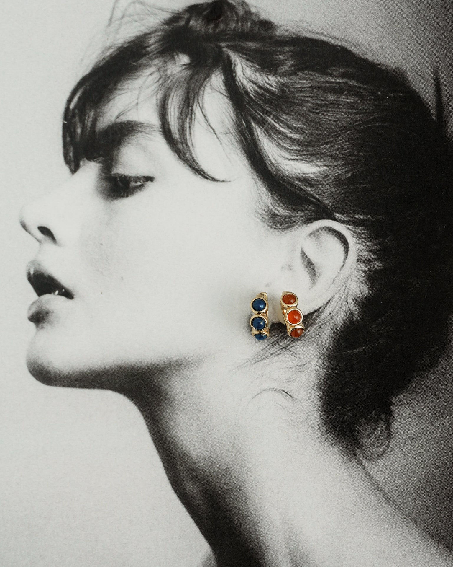 Black and white side profile of a woman wearing Mod + Jo’s Corynn Gemstone Huggies—blue and natural red agate stones set in 18k gold plating. Her dark hair is in a bun, head tilted back to highlight her neck and jawline.