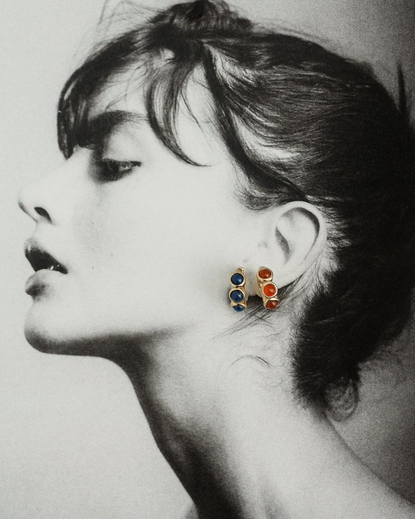 Black and white profile portrait of a woman with her hair tied back. She wears two pairs of stud earrings—one 18k gold plated with blue lapis and one red stone pair—plus Mod + Jo Corynn Gemstone Huggies.