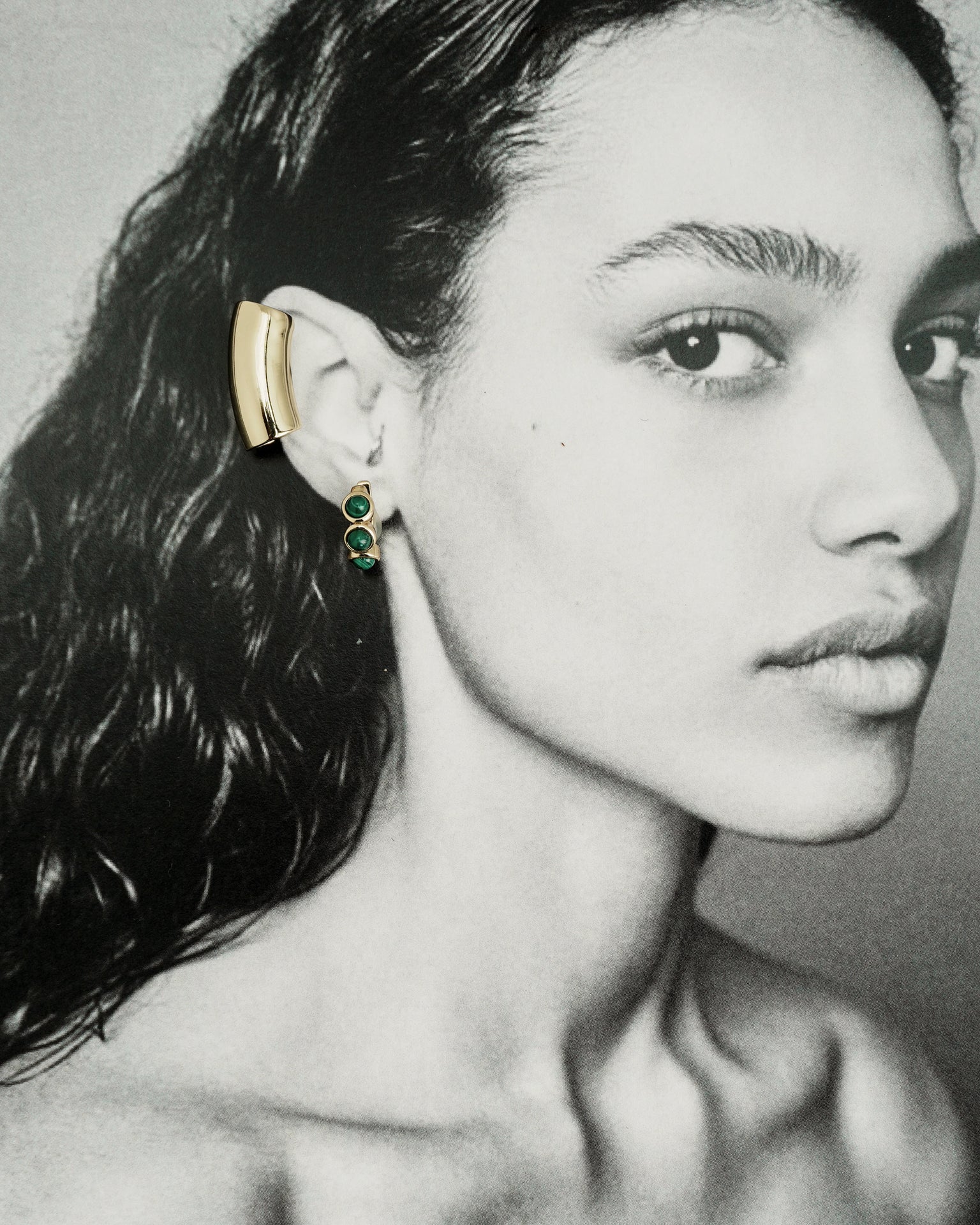 Black and white portrait of a woman with wet, wavy hair, showcasing the Lennon Tube Helix Ear Cuff by Mod + Jo in 18k gold plating and a dangling green gemstone earring. Her face, bare shoulders, and minimalist jewelry style are highlighted.