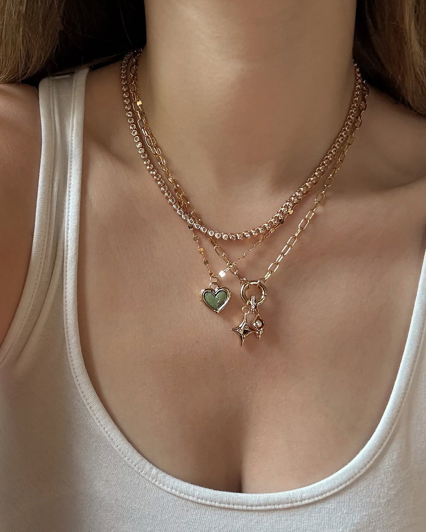 A person in a white tank top layers three gold necklaces, including the Mod + Jo Darcy Charm Clip Necklace, featuring 14k gold plating with a green heart pendant, a star-shaped charm, and one made of small gold links.
