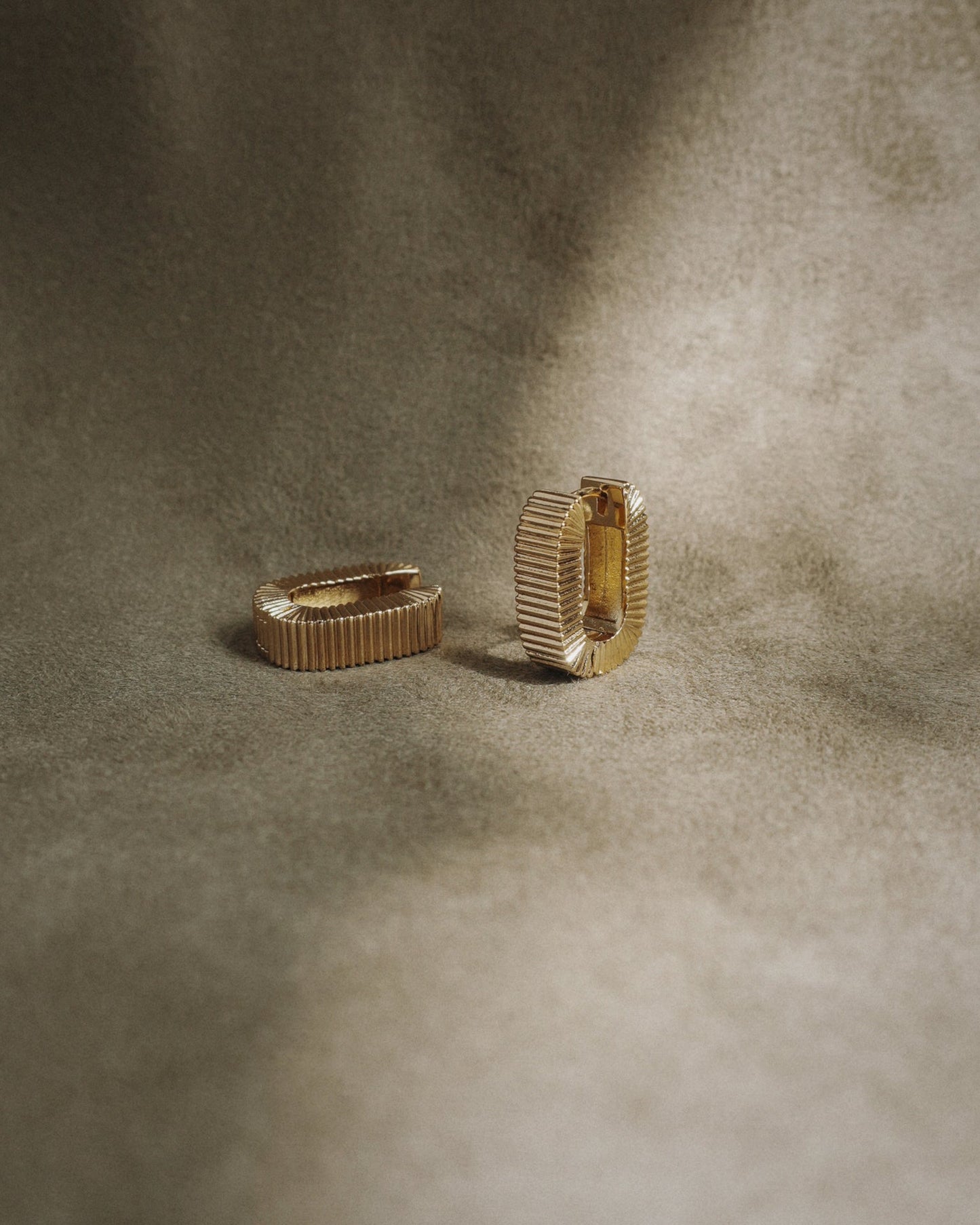 The Elly Fluted Hoops by Mod + Jo, featuring a gold textured design, are displayed on a soft brownish-beige fabric background with gentle lighting.
