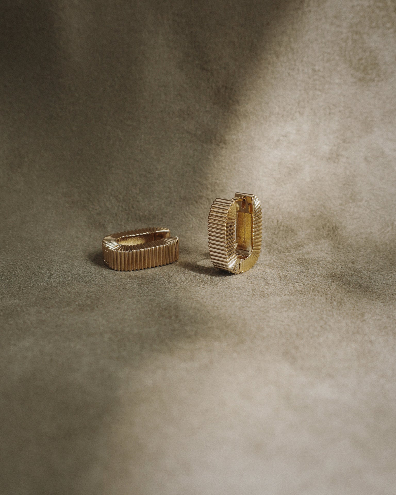 The Elly Fluted Hoops by Mod + Jo, featuring a gold textured design, are displayed on a soft brownish-beige fabric background with gentle lighting.