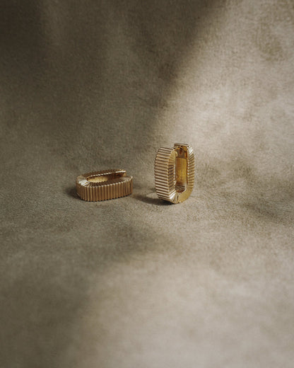 The Elly Fluted Hoops by Mod + Jo, featuring a gold textured design, are displayed on a soft brownish-beige fabric background with gentle lighting.