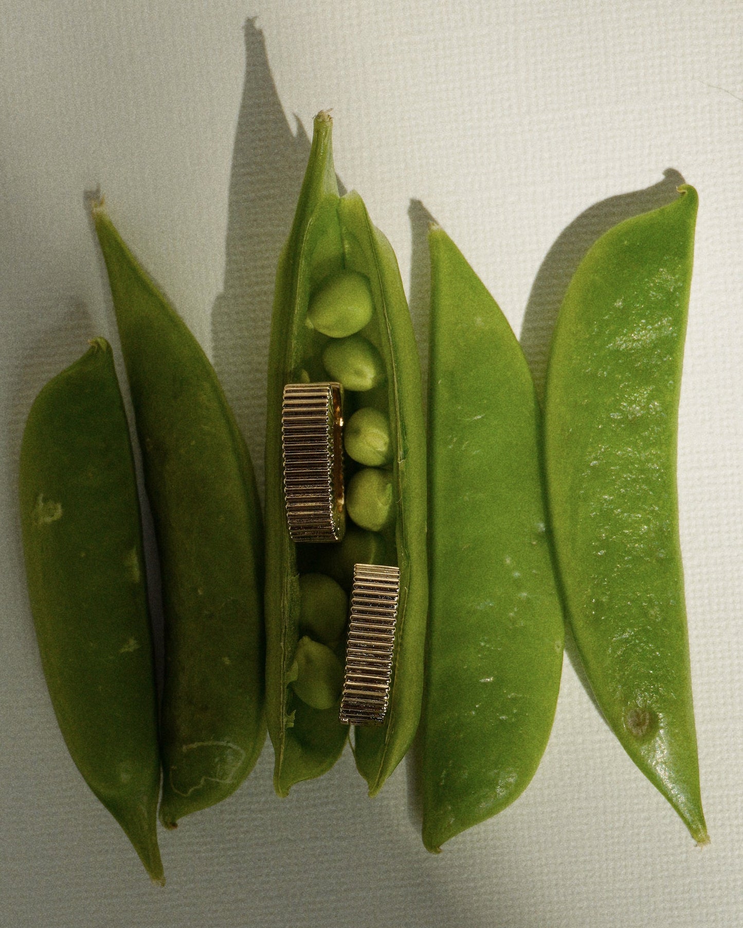 Four green pea pods are arranged on a textured white surface. One pod is open, revealing peas and two small metallic gears inside, creating a striking contrast—just as the Elly Fluted Hoops by Mod + Jo add an unexpected twist to a classic look. Dramatic lighting and shadows complete the scene.