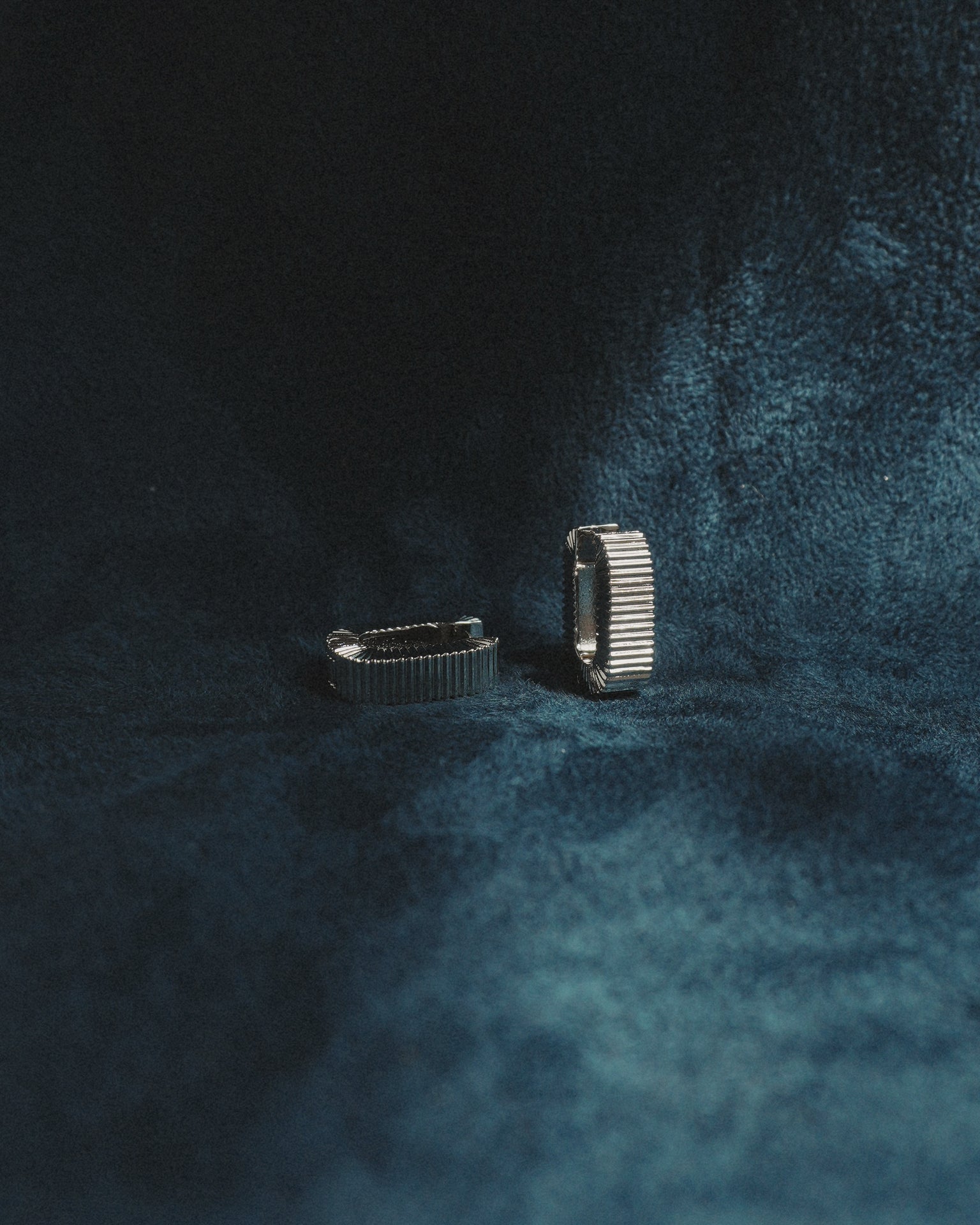A pair of Elly Fluted Hoops by Mod + Jo, crafted in silver, are displayed upright on a dark blue, velvet-textured surface, with soft lighting casting gentle shadows around them.