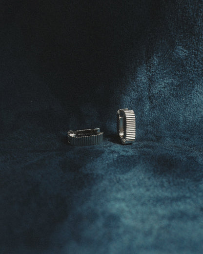 A pair of Elly Fluted Hoops by Mod + Jo, crafted in silver, are displayed upright on a dark blue, velvet-textured surface, with soft lighting casting gentle shadows around them.