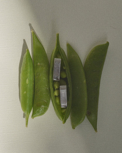 Four green pea pods rest on a light surface; one open pod reveals small peas with a pair of Mod + Jo’s Elly Fluted Hoops nestled inside.
